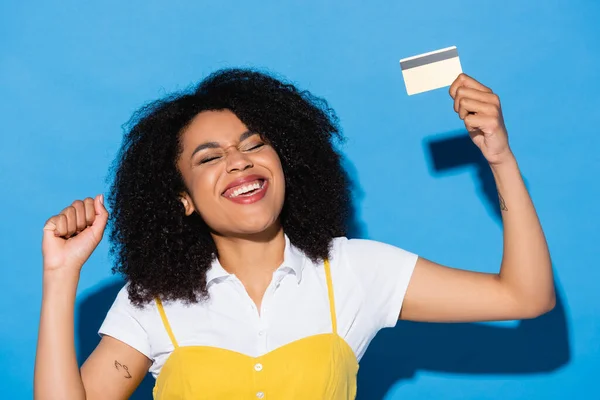 Excited african american woman with closed eyes holding credit card and showing win gesture on blue — Stock Photo