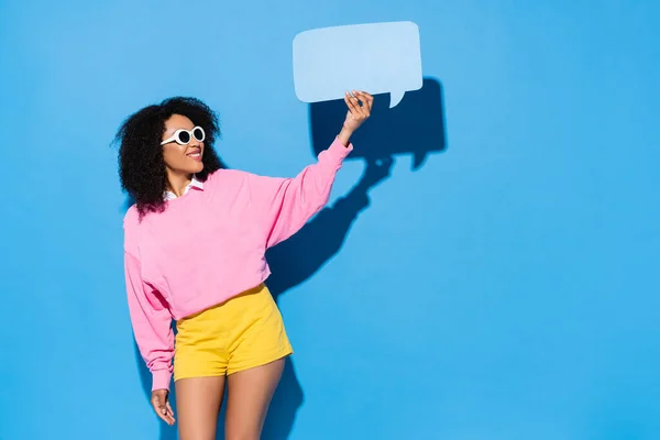 Pleased african american woman in pink sweatshirt and yellow shorts showing empty speech bubble on blue — Stock Photo
