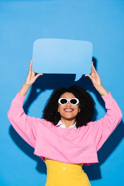 Cheerful african american woman in stylish sunglasses holding blank speech bubble on blue — Stock Photo