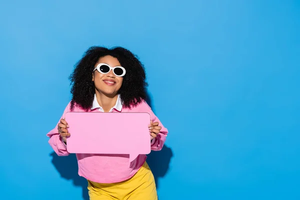 Joyful african american woman in trendy sunglasses and pink sweatshirt holding blank speech bubble on blue — Stock Photo