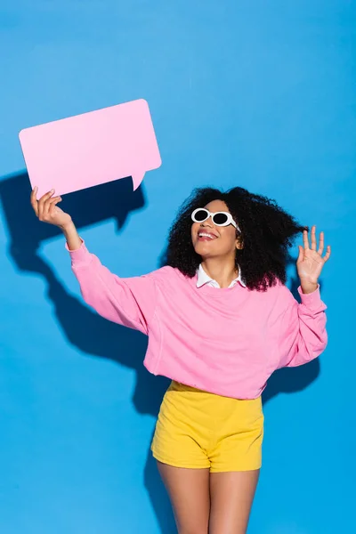 Excited african american woman waving hand while holding speech bubble on blue — Stock Photo