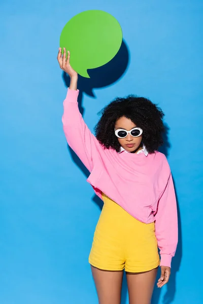 Afro-américaine dans des lunettes de soleil élégantes tenant bulle de pensée vide dans la main levée sur bleu — Photo de stock