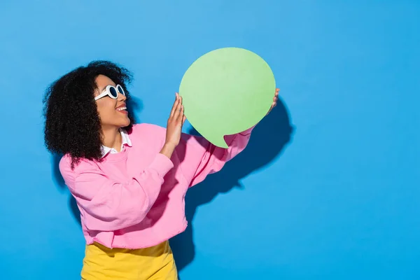 Alegre africana americana mujer en rosa sudadera y gafas de sol celebración en blanco pensamiento burbuja en azul - foto de stock