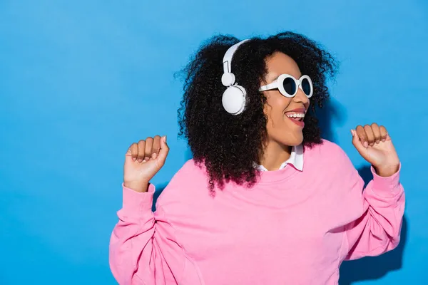 Fröhliche afrikanisch-amerikanische Frau mit stylischer Sonnenbrille, die Musik in Kopfhörern auf blau hört — Stockfoto