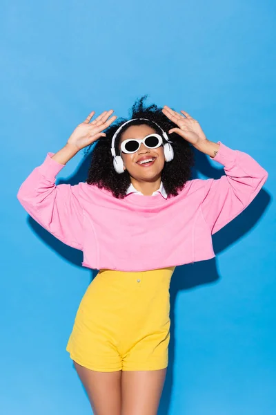 Femme afro-américaine gaie en lunettes de soleil et écouteurs dansant sur bleu — Photo de stock