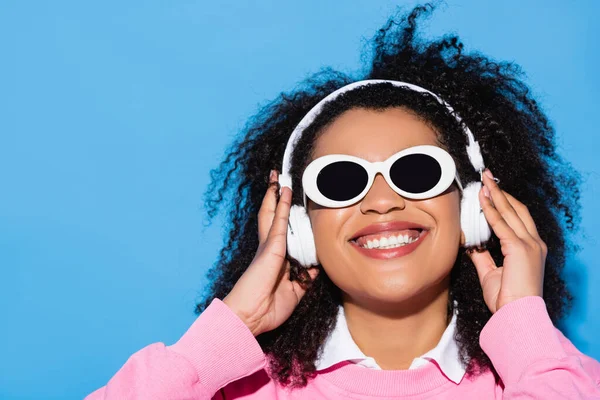 Mujer afroamericana emocionada en gafas de sol de moda tocando auriculares mientras escucha música en azul - foto de stock