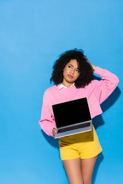 Thoughtful african american woman with laptop touching head and pointing away on blue — Stock Photo