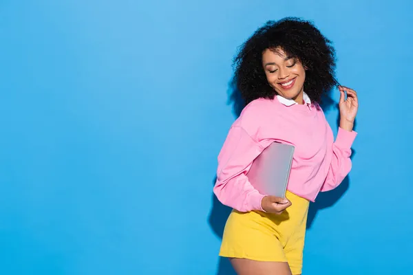 Sorrindo e flertando afro-americano mulher posando com laptop em azul — Fotografia de Stock