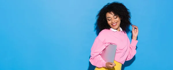 Mujer americana africana coqueta sonriendo mientras posa con el ordenador portátil en azul, bandera - foto de stock