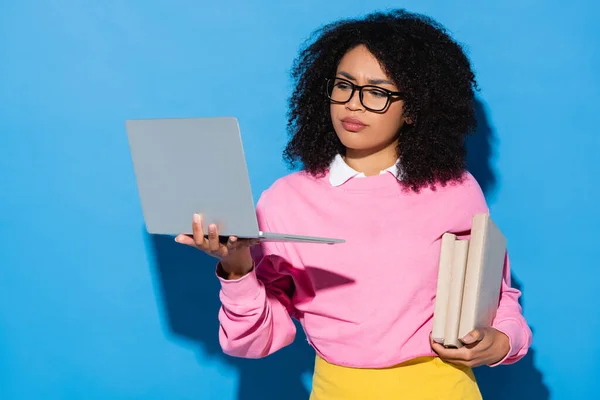 Mujer afroamericana reflexiva y disgustado con libros que miran a la computadora portátil en azul - foto de stock