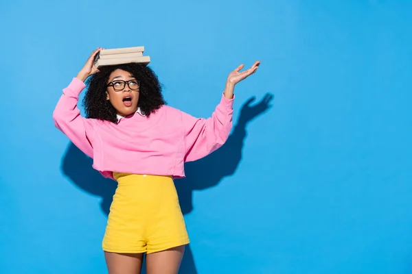 Astonished african american woman with books on head pointing with hand on blue — Stock Photo