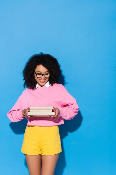 Щаслива афроамериканська жінка в окулярах тримає книги на синьому — стокове фото