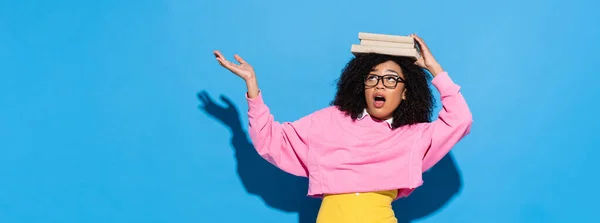 Amazed african american woman with books on head pointing with hand on blue, banner — Stock Photo