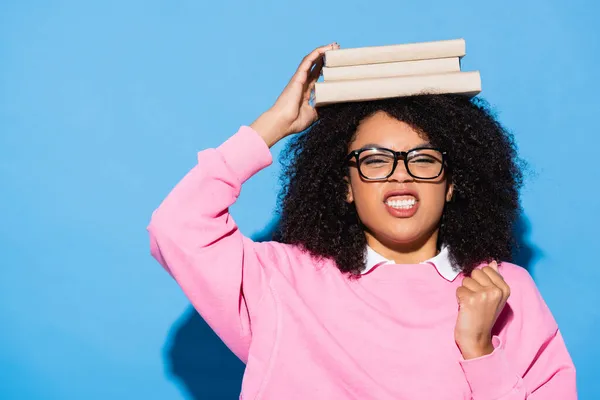 Wütende Afroamerikanerin mit Büchern auf dem Kopf, die Faust auf Blau zeigen — Stockfoto