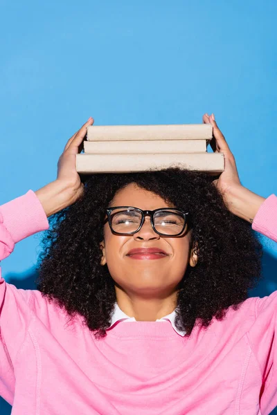 Африканская американка с книгами на голове, хмурящаяся с закрытыми глазами на голубом — стоковое фото