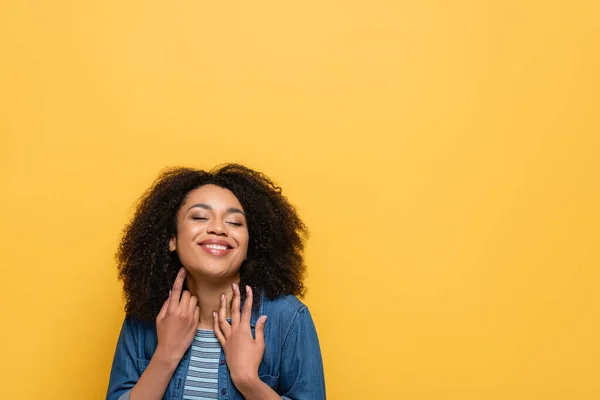 Happy african american woman in denim shirt holding hands near neck while posing isolated on yellow — Stock Photo