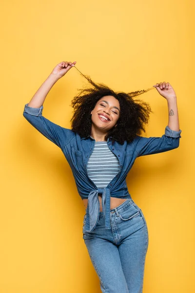 Alegre afroamericana mujer con los ojos cerrados tocando el pelo mientras posando en amarillo - foto de stock