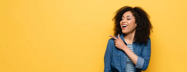 Donna afroamericana ridente che distoglie lo sguardo e punta con il dito isolato sul giallo, striscione — Foto stock