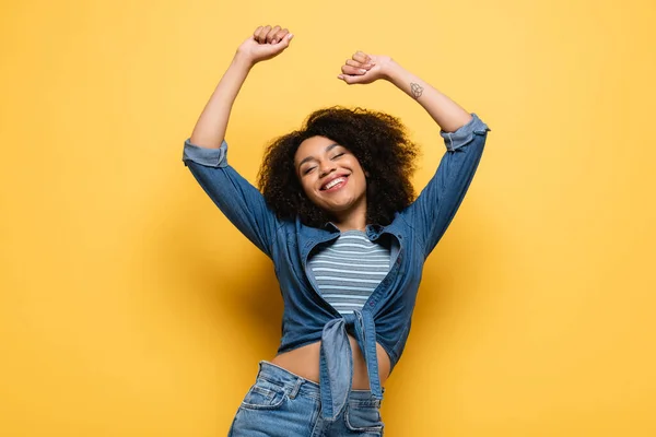 Joyful african american woman in denim shirt dancing with closed eyes and raised hands on yellow — Stock Photo