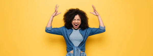 Femme afro-américaine en colère criant avec les yeux fermés et les mains levées sur jaune, bannière — Photo de stock