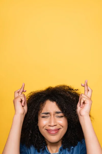 Worried african american woman with closed eyes holding crossed fingers isolated on yellow — Stock Photo