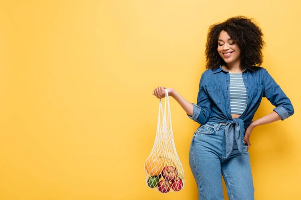 Mulher americana africana alegre segurando saco de cordas com frutas frescas enquanto posando com a mão no quadril no amarelo — Fotografia de Stock