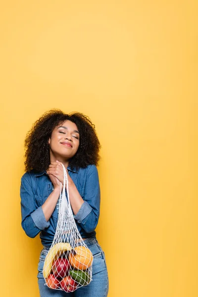 Pleased african american woman with closed eyes holding string bag with ripe fruits isolated on yellow — Stock Photo