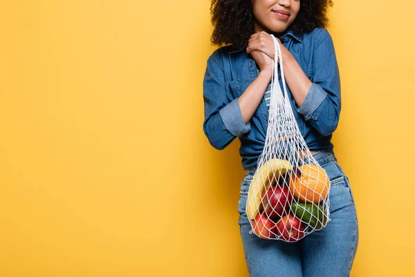 Vista ritagliata di donna afro-americana sorridente che tiene frutti maturi in borsa stringa sul giallo — Foto stock