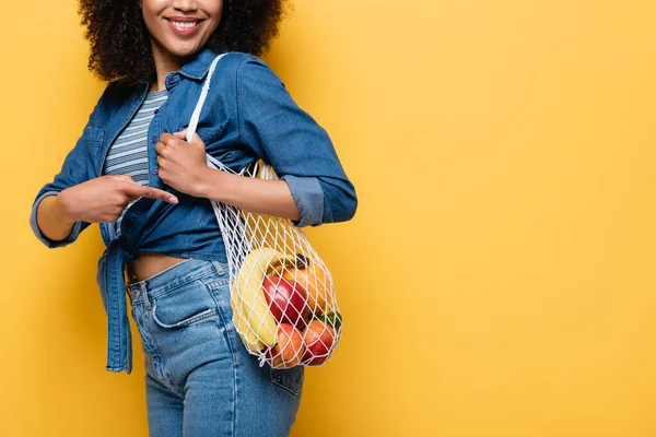 Vista ritagliata di donna afro-americana sorridente che indica borsa stringa con frutti maturi isolati su giallo — Foto stock