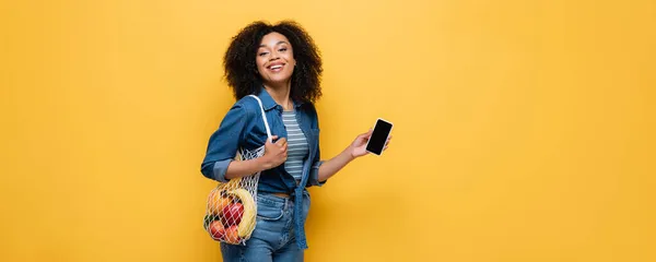 Happy african american woman holding smartphone and string bag with fruits isolated on yellow, banner — Stock Photo