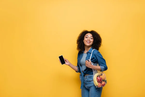 Fröhliche afrikanisch-amerikanische Frau mit Handy und frischen Früchten, die isoliert auf gelb in die Kamera blickt — Stockfoto