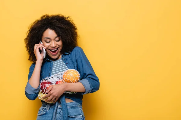 Mujer afroamericana sorprendida con frutas frescas en bolsa de cuerda hablando en el teléfono inteligente en amarillo - foto de stock