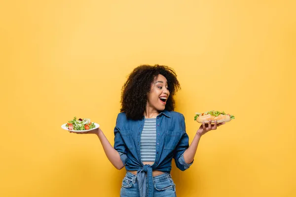 Astonished african american woman looking at hot dog while holding vegetable salad on yellow — Stock Photo