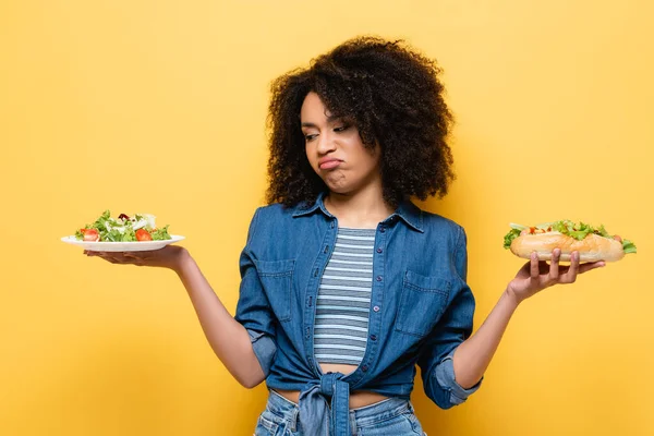 Displeased african american woman looking at vegetable salad while holding hot dog isolated on yellow — Stock Photo
