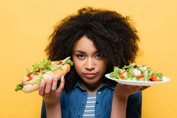 Mujer afroamericana mirando a la cámara mientras sostiene ensalada de verduras frescas y hot dog aislado en amarillo - foto de stock