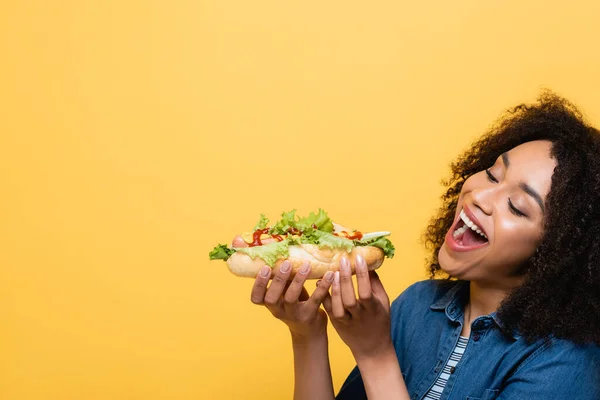 Erfreut afrikanisch-amerikanische Frau essen leckeren Hot Dog isoliert auf gelb — Stockfoto