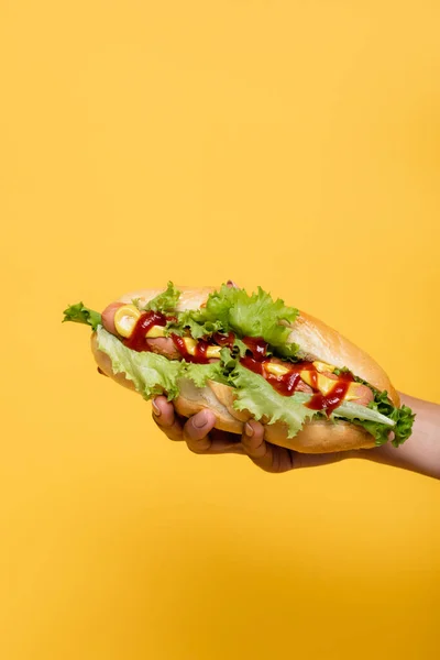 Vista recortada de mujer afroamericana sosteniendo delicioso perrito caliente aislado en amarillo - foto de stock