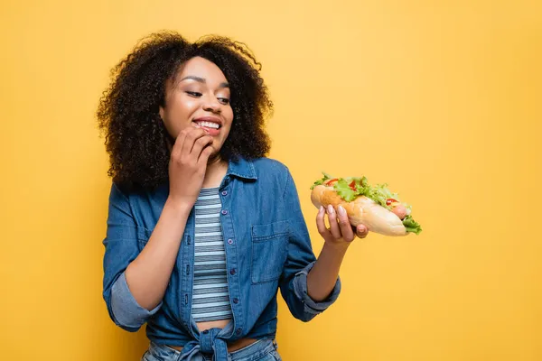 Joyful african american woman looking at delicious hot dog isolated on yellow — Stock Photo