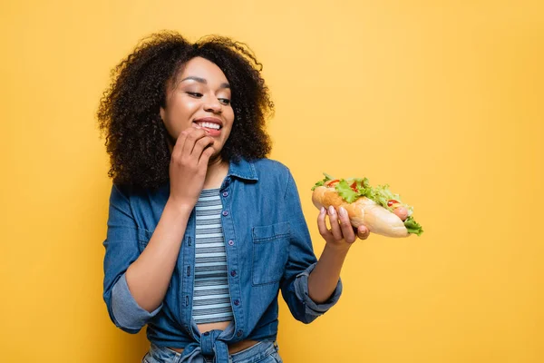 Joyful african american woman looking at delicious hot dog isolated on yellow — Stock Photo