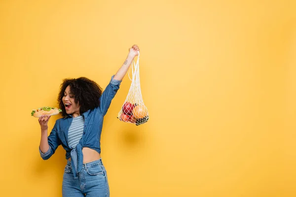 African american woman biting hot dog while holding string bag with fresh fruits in raised hand on yellow — Stock Photo