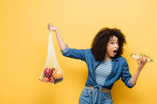 Donna afro-americana con bocca aperta che tiene frutta fresca e hot dog mentre guarda la fotocamera sul giallo — Foto stock