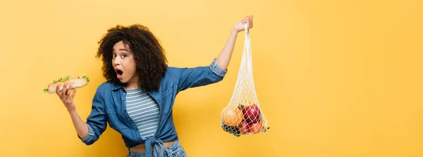 African american woman with open mouth holding fresh fruits and hot dog isolated on yellow, banner — Stock Photo