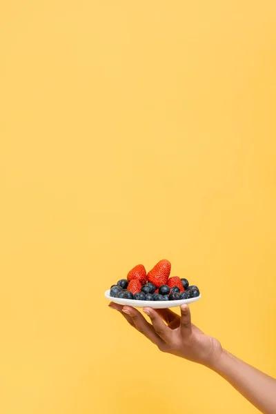 Cropped view of african american woman holding saucer with fresh berries isolated on yellow — Stock Photo