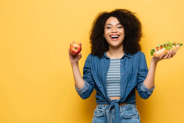 Femme afro-américaine gaie avec pomme fraîche et hot dog regardant caméra sur jaune — Photo de stock