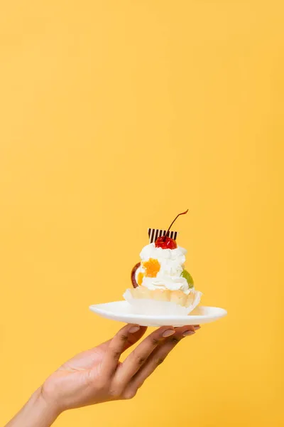 Vista parcial de mujer afroamericana sosteniendo delicioso cupcake con crema batida aislado en amarillo - foto de stock