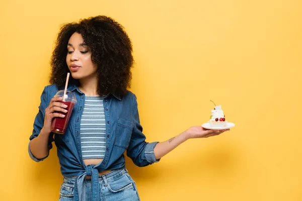 African american woman with tasty cupcake drinking fresh smoothie on yellow — Stock Photo