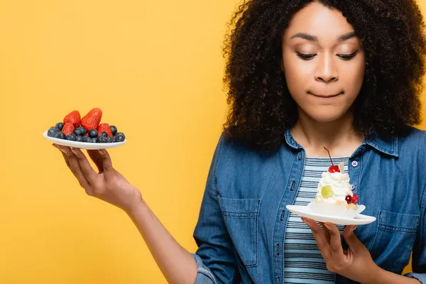 Jovem afro-americana escolhendo entre cupcake e bagas frescas isoladas em amarelo — Fotografia de Stock