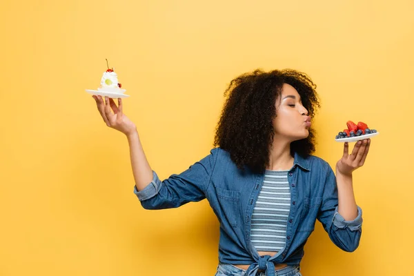 Mujer afroamericana haciendo pucheros labios cerca de bayas frescas mientras sostiene cupcake en amarillo - foto de stock