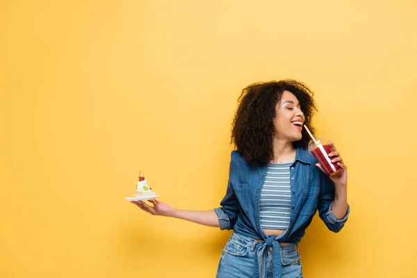 Happy african american woman holding cupcake while drinking fresh smoothie on yellow — Stock Photo