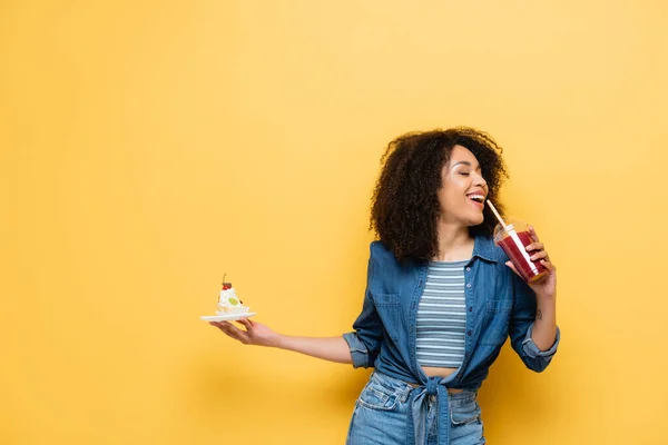 Feliz afro-americana segurando cupcake enquanto bebe smoothie fresco no amarelo — Fotografia de Stock